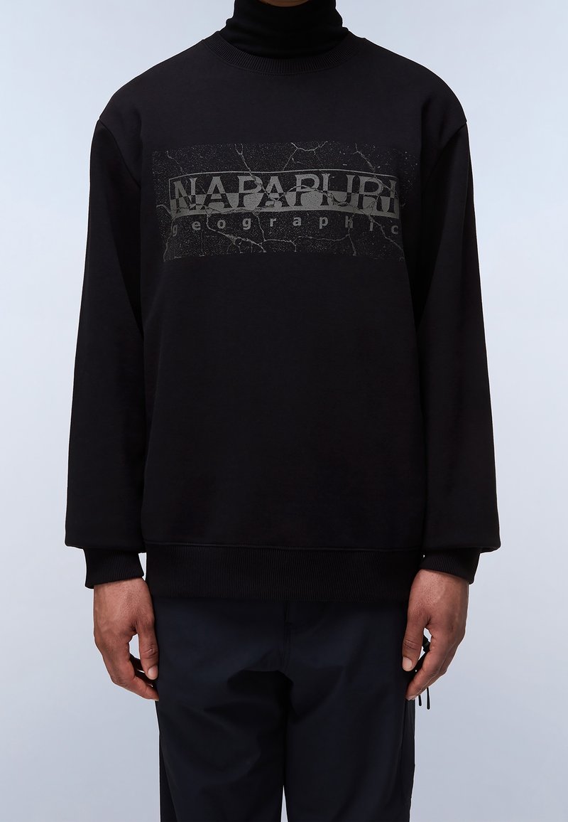 Hombre vistiendo una sudadera negra con el texto "NAPAPIJRI geographic" y un patrón agrietado en el pecho, combinada con pantalones negros.