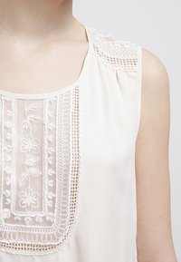 Blouse sans manches crème avec des détails en dentelle autour de l'encolure et des motifs floraux brodés sur le panneau avant. Tissu à texture lisse.