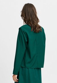 Chemise verte à col rabattu, à manches longues, en tissu lisse. Elle présente une couture au dos et une coupe carrée qui descend jusqu'à la taille.