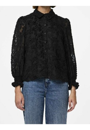 Vrouw draagt een zwarte doorzichtige blouse met bloemmotief, lange mouwen met geplooide manchetten en een blauwe spijkerbroek, staand tegen een witte achtergrond.