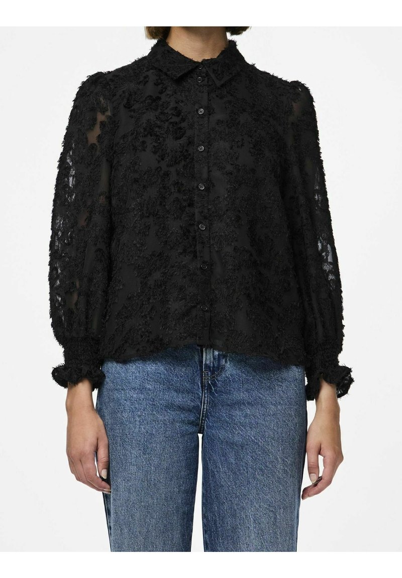 Femme portant un chemisier noir transparent à texture florale avec manches longues et poignets volantés, et un jean en denim bleu, debout sur fond blanc.