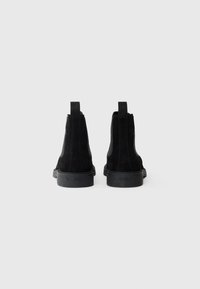 Cizme negre din suede la glezna, cu un talpă din cauciuc neted, panouri elastice pe laterale și o curea la spate. Design minimalist, linii curate.