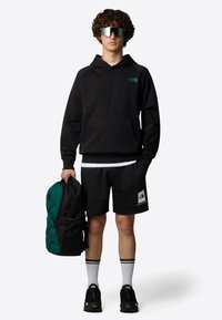 Sudadera negra con bolsillo frontal, combinada con pantalones cortos negros. Gafas de sol espejadas plateadas y una mochila verde-negra. Zapatos negros con suelas texturizadas.