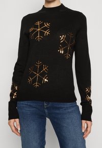 Pull noir en tricot avec manches longues, décoré de flocons de neige métalliques dorés sur le devant et les manches. Col et poignets côtelés.