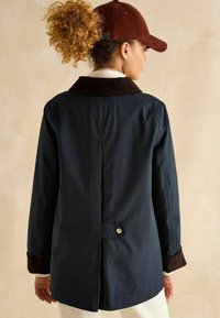 Jachetă impermeabilă navy cu guler și manșete din catifea stacojie, având o ventilație pe spate, detalii cu nasturi aurii și o croială relaxată.