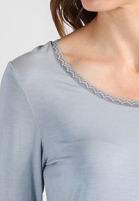 Vaaleanharmaa paita, jossa on sileä pinta, pyöreä kaula-aukko ja koristeellinen pitsireunus kauluksen reunassa. Pitkät hihat.
