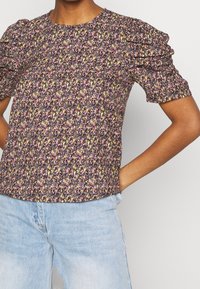 Femme portant un haut à manches courtes en fleurs avec des volants, orné de petites fleurs violettes, jaunes et blanches, associé à un jean bleu clair.