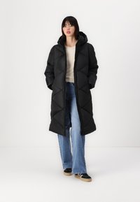 Black puffer coat with a hood, featuring a zip closure, diagonal quilted pattern, and side pockets, worn over light blue jeans and black shoes.