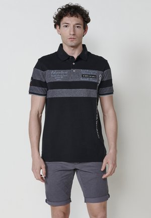 Schwarzes Poloshirt mit grauen horizontalen Streifen, mit textlichen Details. Kurze Ärmel, kontrastierender Kragen mit Knöpfen und schmale Passform, kombiniert mit grauen Shorts.