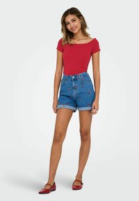 Röd kortärmad topp i kombination med blå denimshorts med blommönster. Modellen har på sig röda balettskor och står på en ljus bakgrund.