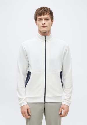 Junger Mann mit kurzen braunen Haaren, trägt eine weiße Jacke mit Reißverschluss, marineblaue Seitenpaneele und hellgraue Hose vor einem schlichten Hintergrund.