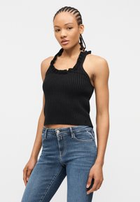 Γυναίκα με πλεγμένα μαλλιά, που φορεί μαύρο ριπ τοπ χωρίς μανίκια και μπλε τζιν, στέκεται μπροστά σε ένα απλό λευκό φόντο.