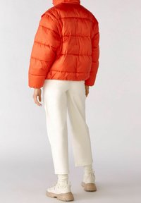 Doudoune orange vif, présentant un design matelassé, associée à un pantalon blanc et des baskets beiges. Le matériau semble brillant.