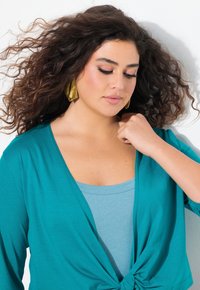 Cardigan en tricot turquoise avec un détail de nœud sur le devant, porté par-dessus un débardeur bleu clair. Des boucles d'oreilles géométriques dorées complètent la tenue.