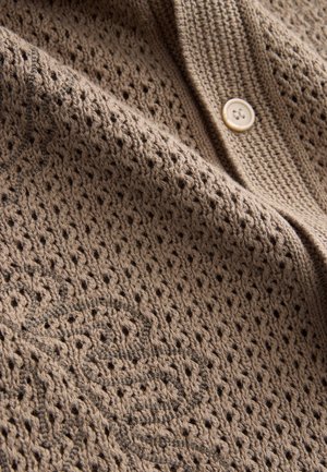 Bruine gebreide stof met een gestructureerd, open geweven patroon. Voorzien van een lichtgekleurde knoop en geborduurde details in een donkere draad.