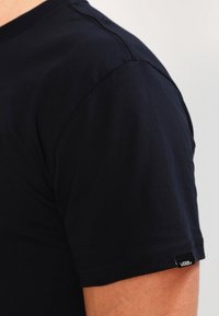 Zwarte Vans t-shirt met korte mouwen, gedragen op een arm met zichtbare stiksels en een klein logo-label op de mouwzoom.