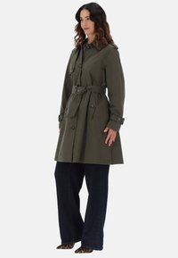 Trench coat verde oliva con cintura rialzata, dettagli a bottoni e spalline, abbinato a jeans scuri a gamba larga e scarpe con stampa leopardata.