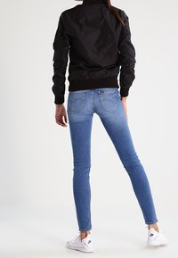 Zwarte bomberjack met geribbelde manchetten, gecombineerd met lichtblauwe skinny jeans. Model draagt witte sneakers, gezien vanaf de achterkant.