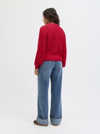 Röd stickad tröja med ett texturerat mönster, ribbad krage och lös passform, i kombination med ljusblå vida jeans och bruna skor.