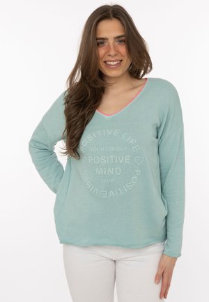 Junge Frau mit langen braunen Haaren trägt einen hellblauen Pullover mit dem Schriftzug „Positives Leben, positiver Geist, gute Energie“ und lächelt in die Kamera.