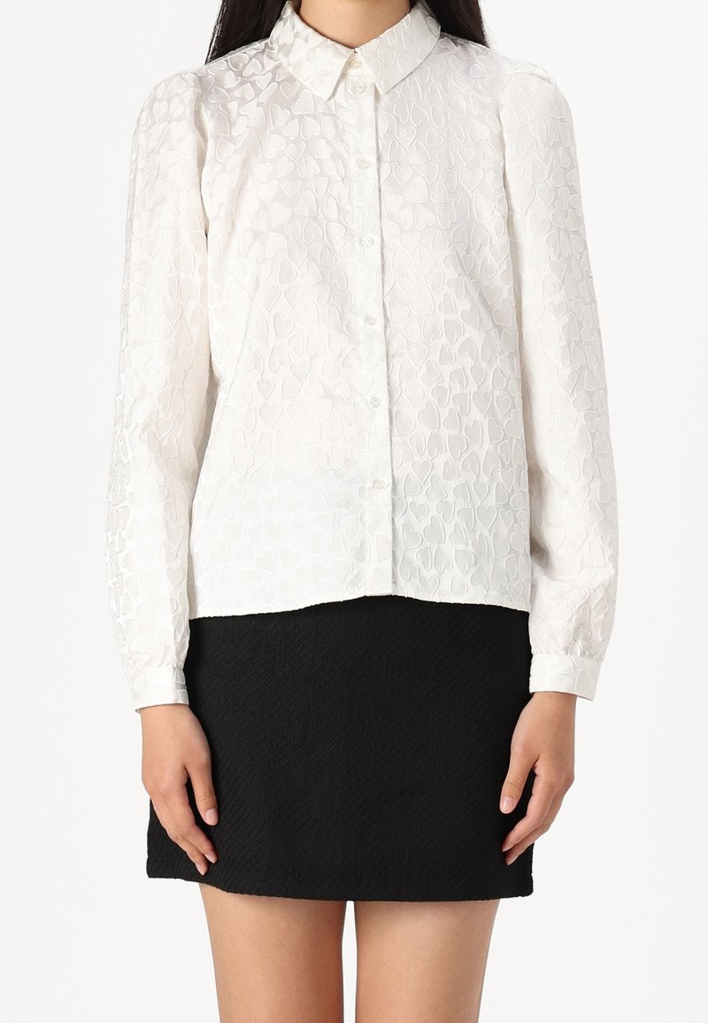Chemise blanche à manches longues avec un motif cœur texturé, col à revers et boutonnée, associée à une jupe noire texturée.
