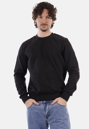 Uomo con capelli ricci e barba chiara, indossa una felpa nera e jeans blu, in piedi con le mani nelle tasche su sfondo bianco.