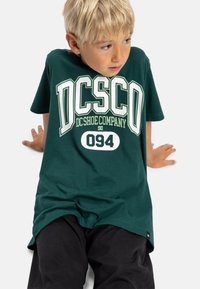 Zelené bavlněné tričko s potiskem "DC Shoe Company" v bílé a světle zelené barvě. Tričko má volný střih, kulatý výstřih a krátké rukávy.