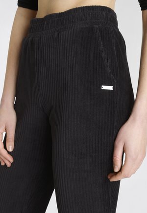 Black corduroy pants with vertical ribbing, elastic waistband, side pockets, and a small metallic accent on the front for detail.
