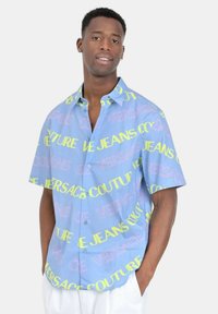 Camicia azzurra con maniche corte e bottoni, caratterizzata da grandi grafiche testuali colorate in rosa e verde, realizzata in tessuto leggero.