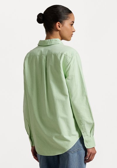 Chemise à manches longues vert clair avec un col, un pli au dos et des poignets boutonnés, associée à un jean en denim bleu.