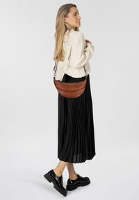 Borsa a tracolla in pelle marrone con design curvo, cuciture dettagliate e una lunga tracolla regolabile, abbinata a una gonna nera plissettata.