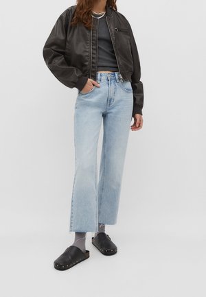 Vrouw die een zwarte bomberjas draagt, een zwart cropped topje, lichtblauwe high-waisted wijd uitlopende spijkerbroek, grijze sokken en zwarte slip-on klompen.
