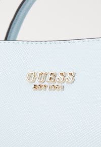 Borsa sintetica azzurro chiaro con superficie testurizzata, dotata di un logo dorato che recita "GUESS EST 1981" ben visibile.
