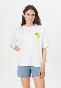 Camiseta de algodón blanca con mangas cortas, que presenta un gráfico de un limón amarillo y una hoja verde en el área del pecho izquierdo. Combinada con pantalones cortos de mezclilla.