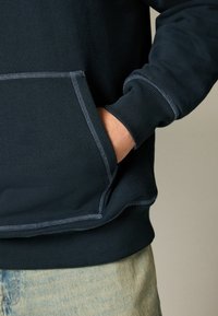 Sudadera azul marino con un bolsillo frontal, puños acanalados y costuras acabadas resaltadas por un hilo en contraste de color azul claro. La tela es suave y texturizada.