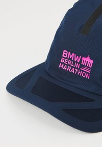 Tamsiai mėlyna bėgimo kepurė su tinklinėmis dalimis, su rožiniu siuvinėtų tekstu: "BMW BERLIN MARATHON" ir vertikaliu juodu raštu priekyje.