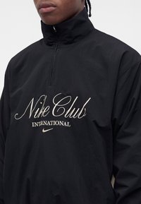 Άτομο που φοράει ένα μαύρο πουλόβερ Nike Club International με φερμουάρ στο μέρος του στήθους και κεντημένο λευκό λογότυπο στο στήθος.