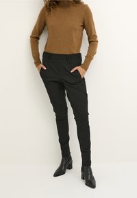 Pull col roulé marron ajusté et pantalon noir fuselé avec ceinture élastique. Le modèle porte des bottines à talons noires. Texture lisse.