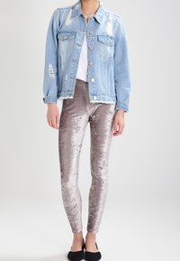 Kvinna klädd i en ljusblå slitna denimjacka, vit skjorta, silverfärgade krossad sammet leggings och svarta lägenheter, stående mot en enkel bakgrund.