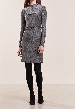 Kvinde iført en tætsiddende metallic grå langærmet kjole med flæsedetaljer, kombineret med sorte ruskindsstøvler, der går til låret, stående foran en ensfarvet baggrund.