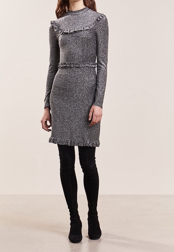 Femme portant une robe longue moulante gris métallisé à manches longues avec des détails à volants, associée à des bottes hautes en daim noir, debout sur un fond uni.