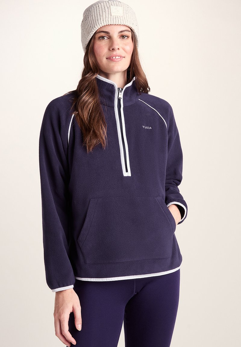 Young woman wearing a navy blue fleece pullover with white trim and a beige knit beanie, standing against a plain background.