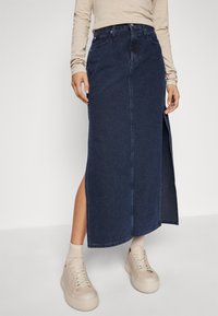 Denimmaxiklänning i mörkblått med hög slits. Designad med en åtsittande midja och mjukt, texturerat tyg. Kombinerad med ljusa sneakers.