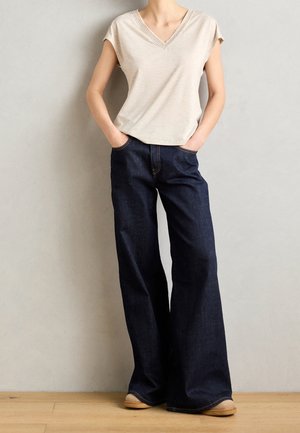 Personne portant un haut beige à manches courtes et col en V, ainsi qu'un jean large foncé, debout les mains dans les poches contre un mur uni.