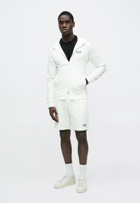 Bela jopa z zadrgo in kratke hlače iz mehkega materiala z logotipom EA7. Obleko dopolnjujejo superge in bele nogavice.