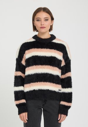 Jeune femme aux cheveux courts portant un pull rayé duveteux noir, blanc et pêche et un pantalon foncé, debout devant un fond uni.