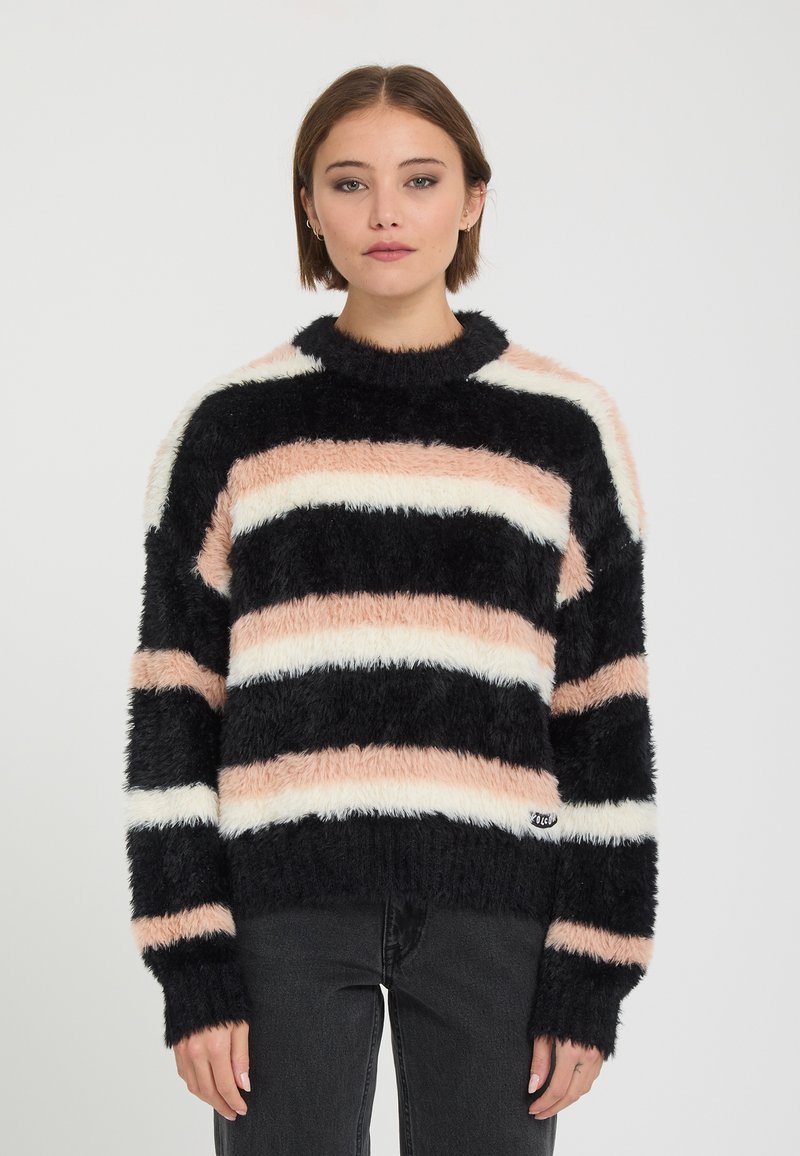 Jeune femme aux cheveux courts portant un pull rayé duveteux noir, blanc et pêche et un pantalon foncé, debout devant un fond uni.
