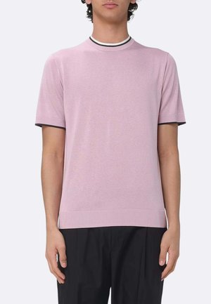 Uomo che indossa un maglione leggero rosa chiaro a maniche corte con collo a righe nere e bianche, abbinato a pantaloni neri, in piedi contro uno sfondo semplice.