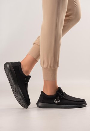 Füße in schwarzen Slip-on-Schuhen mit dicken Sohlen und Kordelzügen, kombiniert mit beigen Jogginghosen vor einem neutralen Hintergrund.