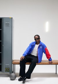 Blue and red windbreaker jacket with logo, white T-shirt, black sweatpants, seated on a wooden bench next to a gray locker and a basketball.